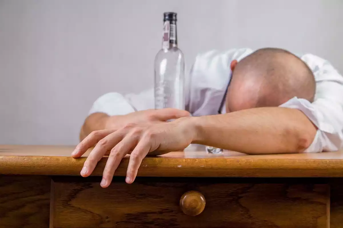 Hombre con la cabeza agachada sobre la mesa con una botella de alcohol vacía en la mano