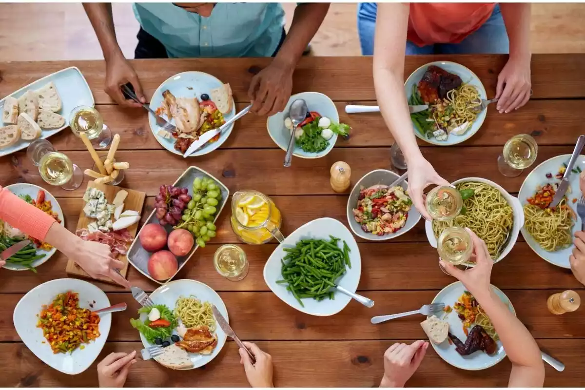 Personas comiendo en una mesa grande mucha comida con verduras, frutas y legumbres