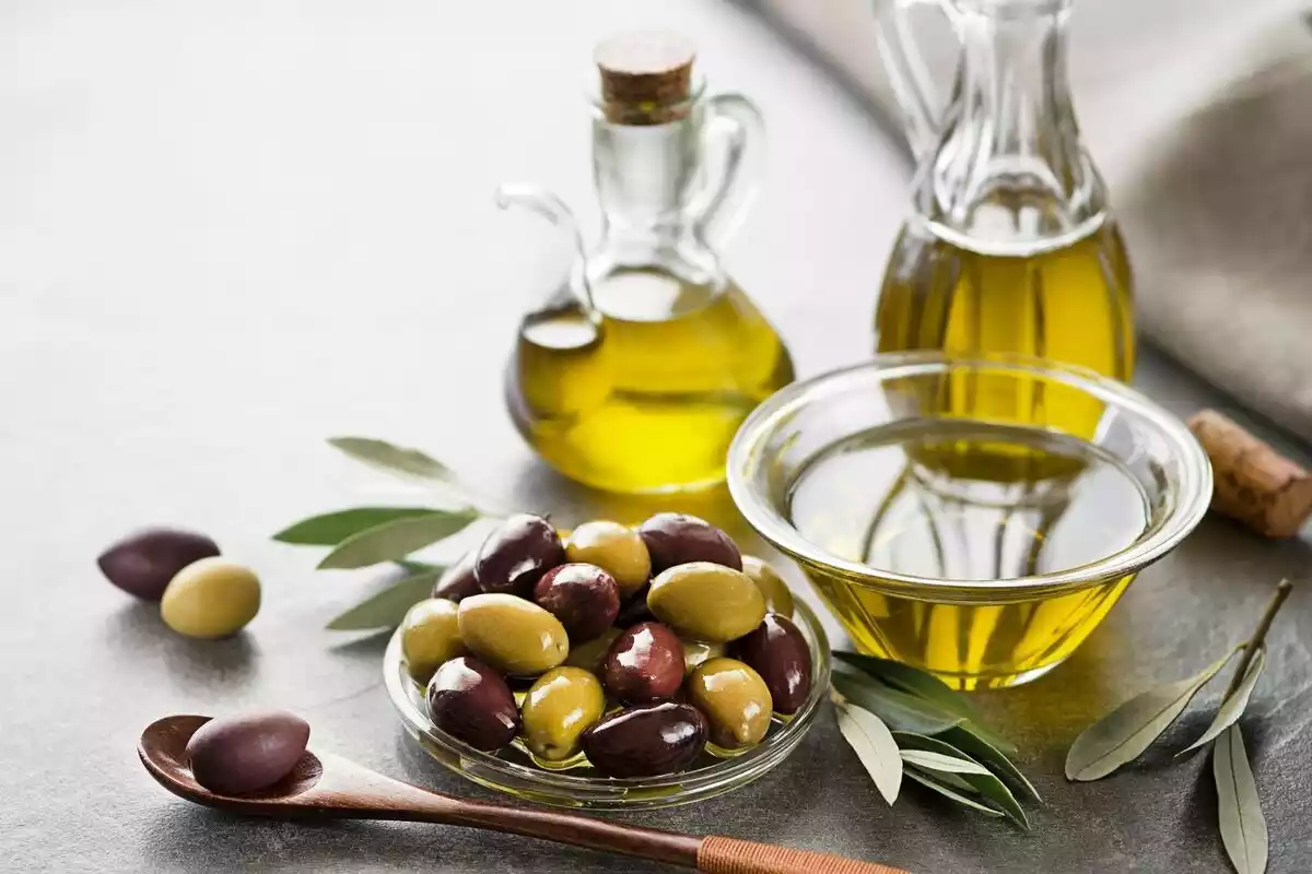 Aceite de oliva en un vaso y dos aceiteras junto a un plato lleno de aceitunas y una cuchara