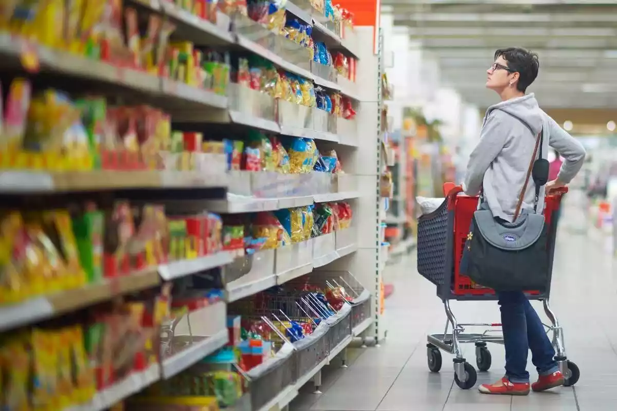 Mujer en el pasillo de un supermercado con un carro de la compra mirando una estantería llena de fideos