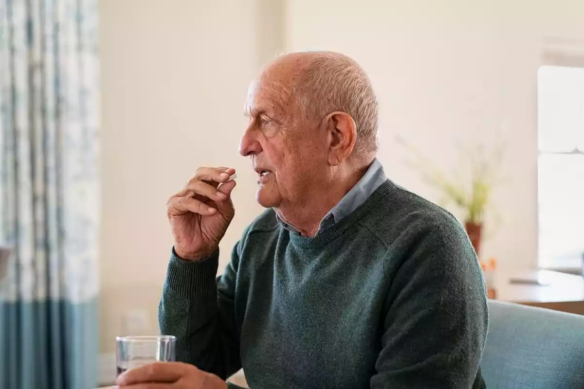 Un señor mayor medicándose con una pastilla mientras sujeta un vaso de agua