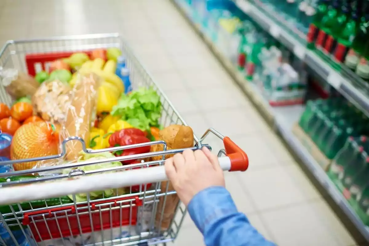 Un carrito de supermercado lleno con frutas y verduras y una mano aguantándolo