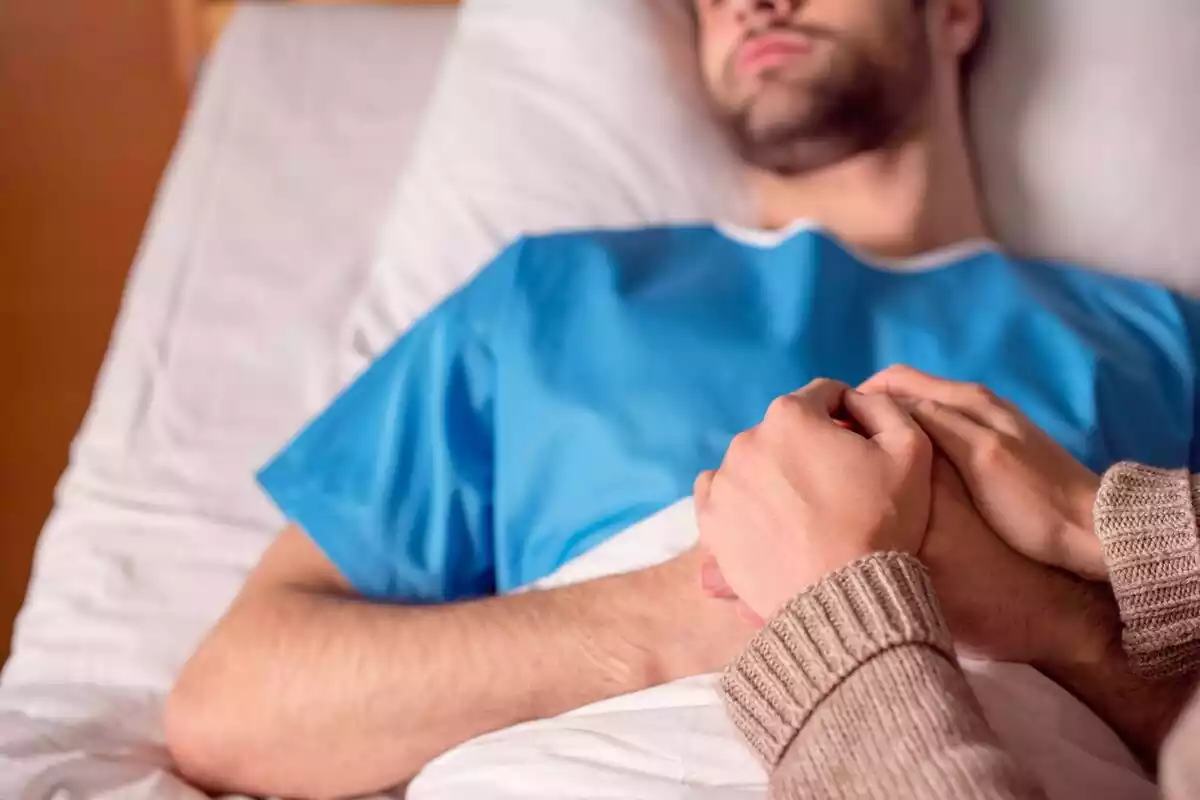 Persona sosteniéndole la mano a un hombre que está en una camilla de hospital