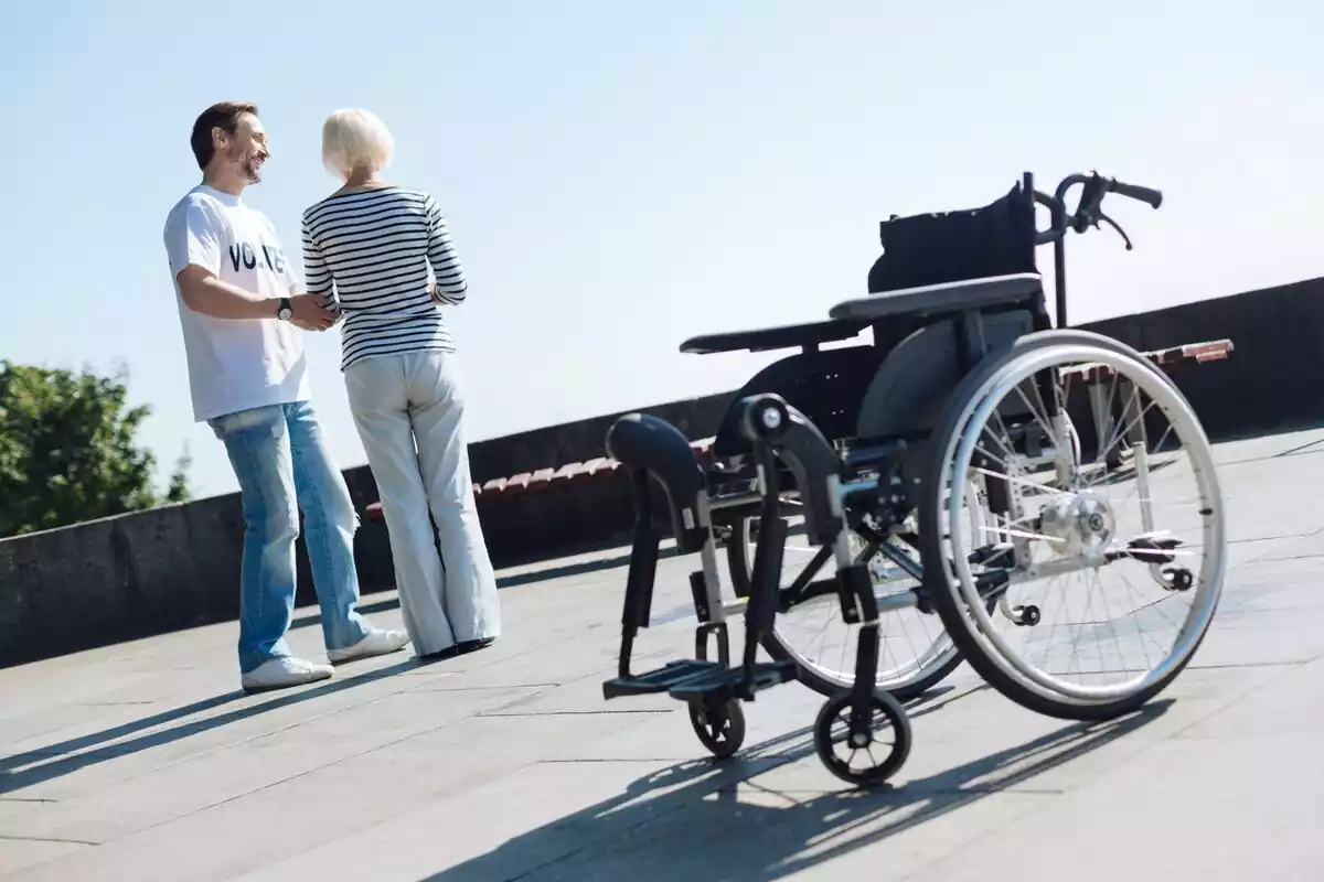 Mujer mayor con un hombre que le acompaña y una silla de ruedas de fondo