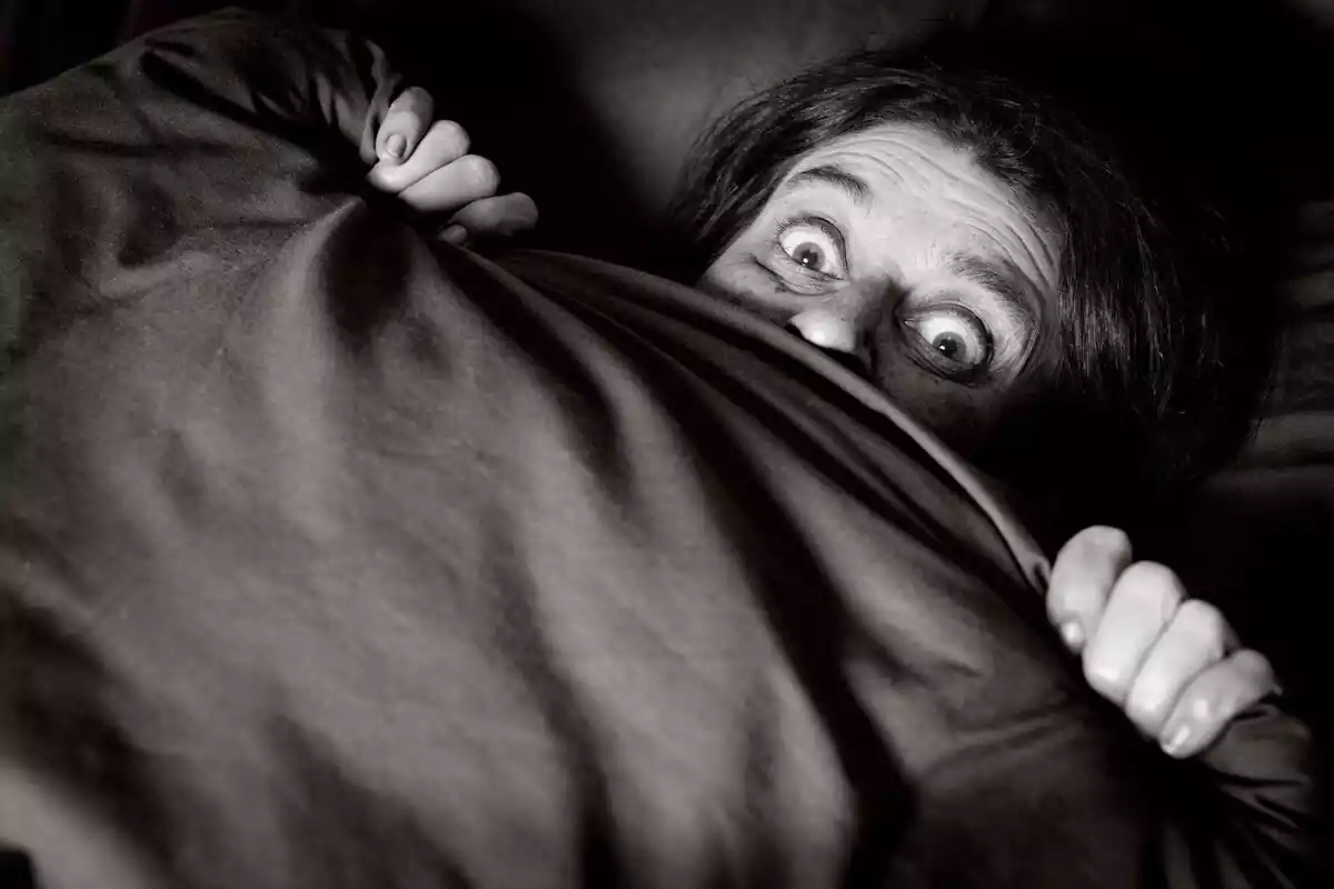 Persona acostada en una cama y tapada hasta la nariz con expresión de pánico
