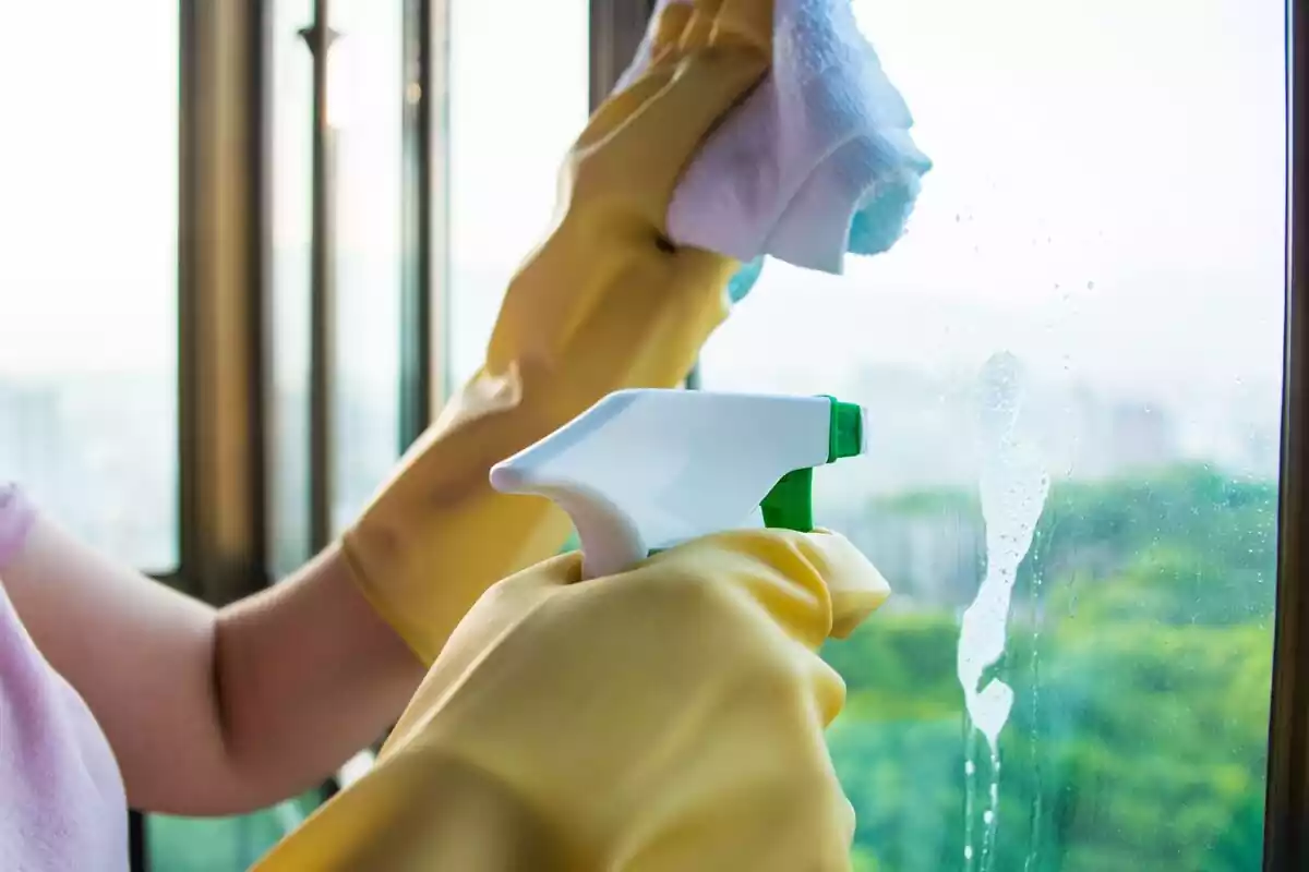 Primer plano de unas manos limpiando el cristal de una ventana con guantes, bayeta y pulverizador