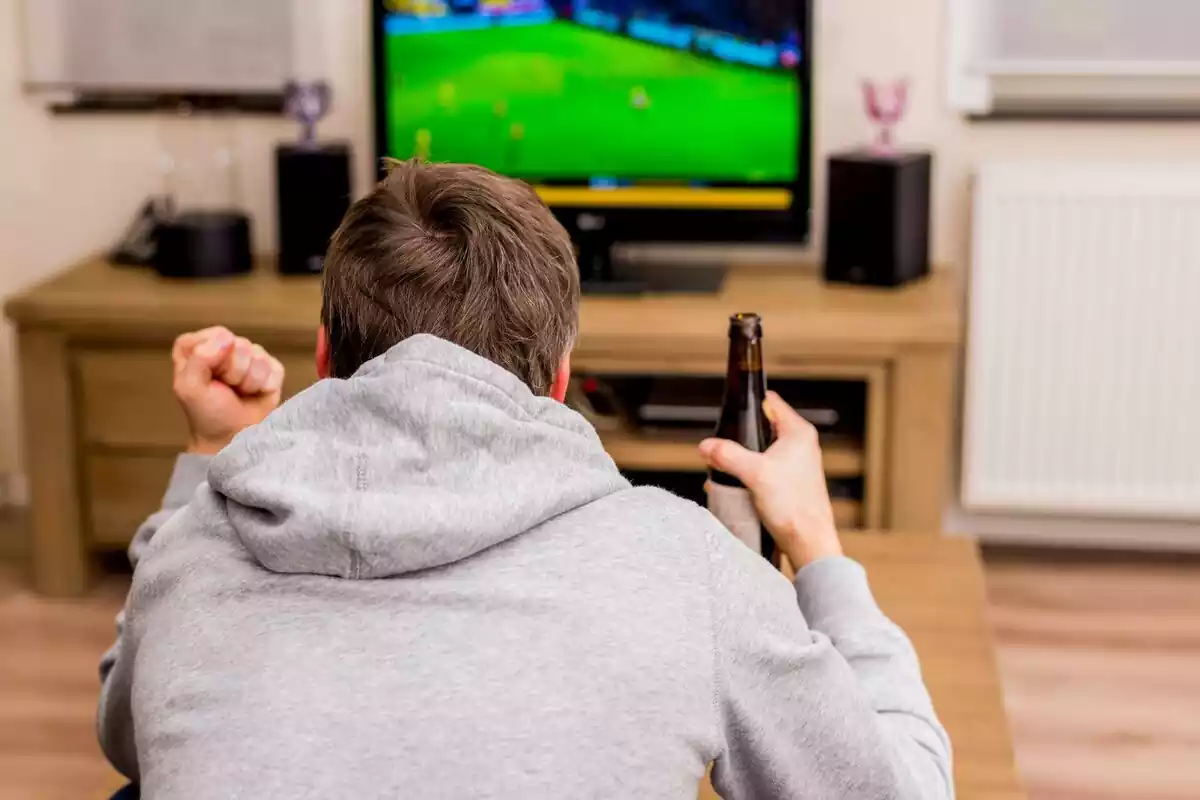Un hombre con sudadera gris viendo un partido de futbol en la televisión