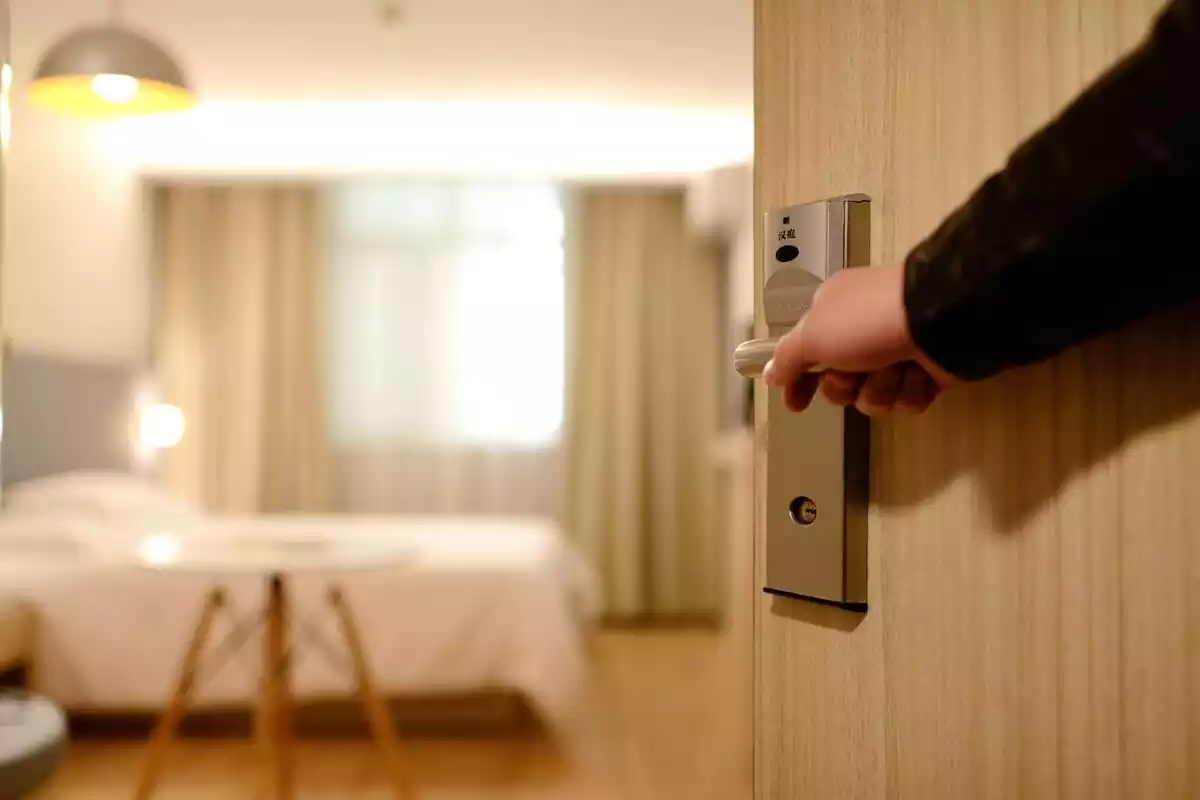 Una mano abriendo la puerta de la habitación de un hotel con la cama detrás