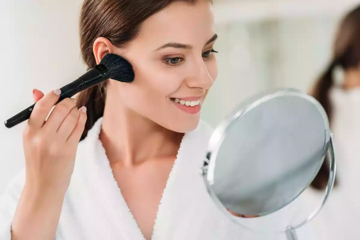 Mujer poniéndose bronceador sobre la mejilla con una brocha mientras se mira al espejo