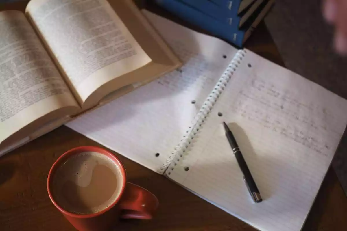 Una taza de café, un libro y una libreta con un bolígrafo y apuntes