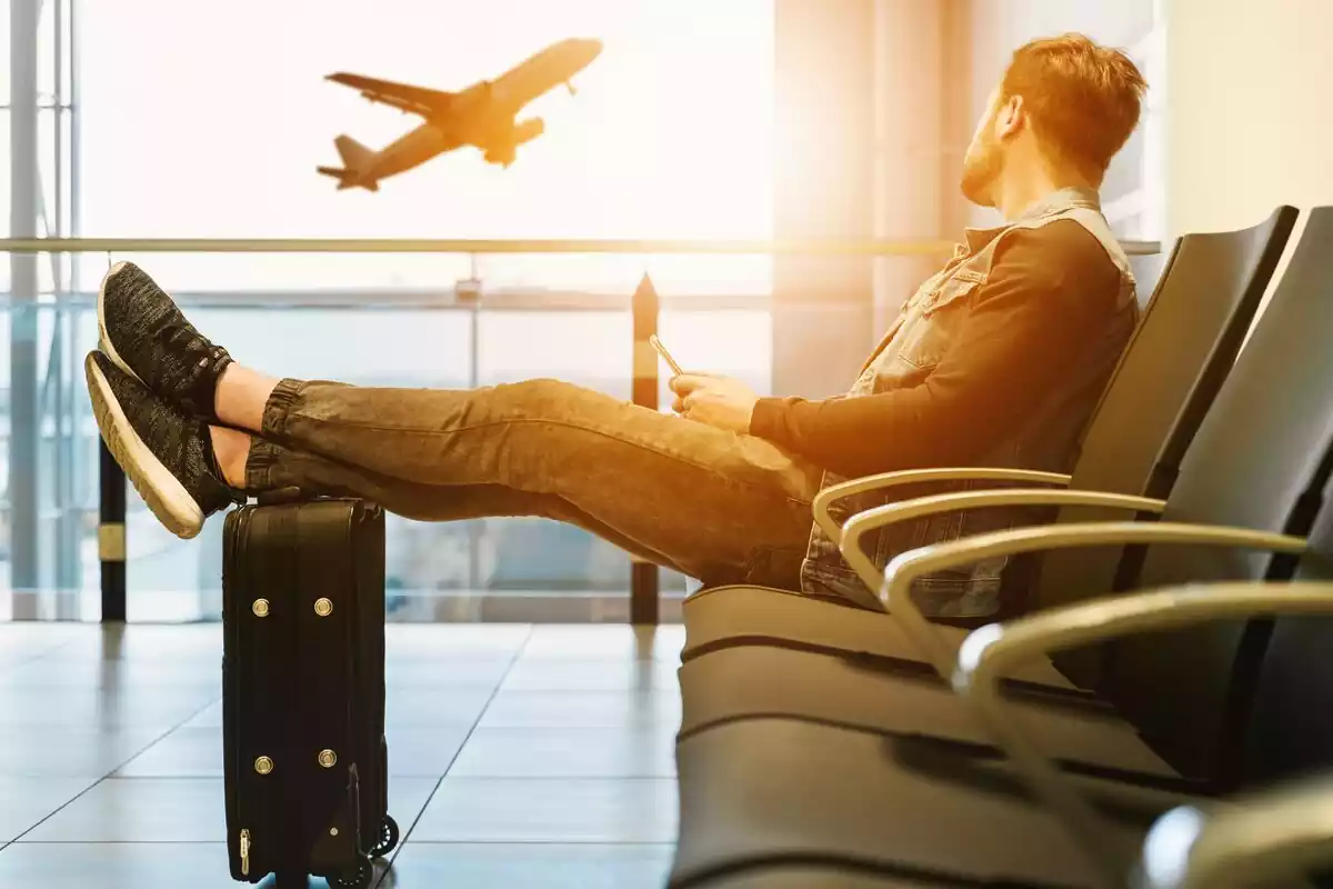 Hombre sentado en un aeropuerto con su maleta al lado, viendo despegar un avión desde la pista