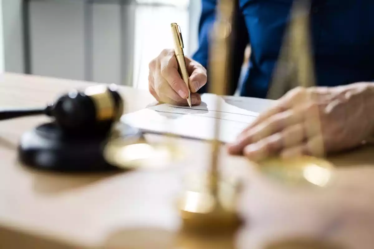 Persona firmando un documento en un juzgado con un mazo y una balanza sobre la mesa