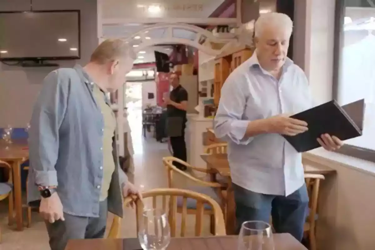 Dos personas están en un restaurante, una de ellas sostiene un menú mientras la otra se prepara para sentarse. Dos personas están en un restaurante, una de ellas sostiene un menú mientras la otra se prepara para sentarse.