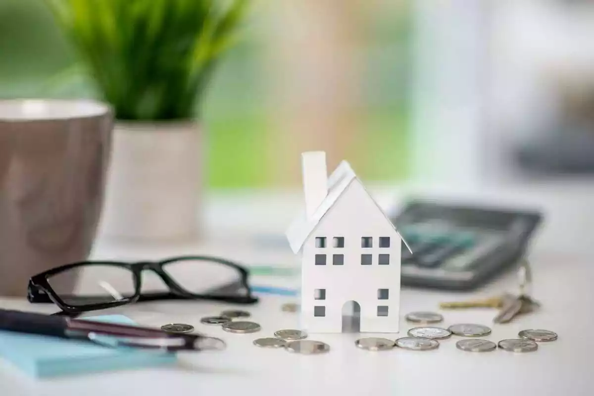 Imagen de una maqueta de una casa en miniatura con varias monedas y unas gafas y una calculadora alrededor