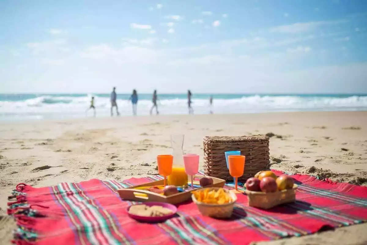 Comida y zumo de naranja sobre un mantel en la playa con varias personas a lo lejos