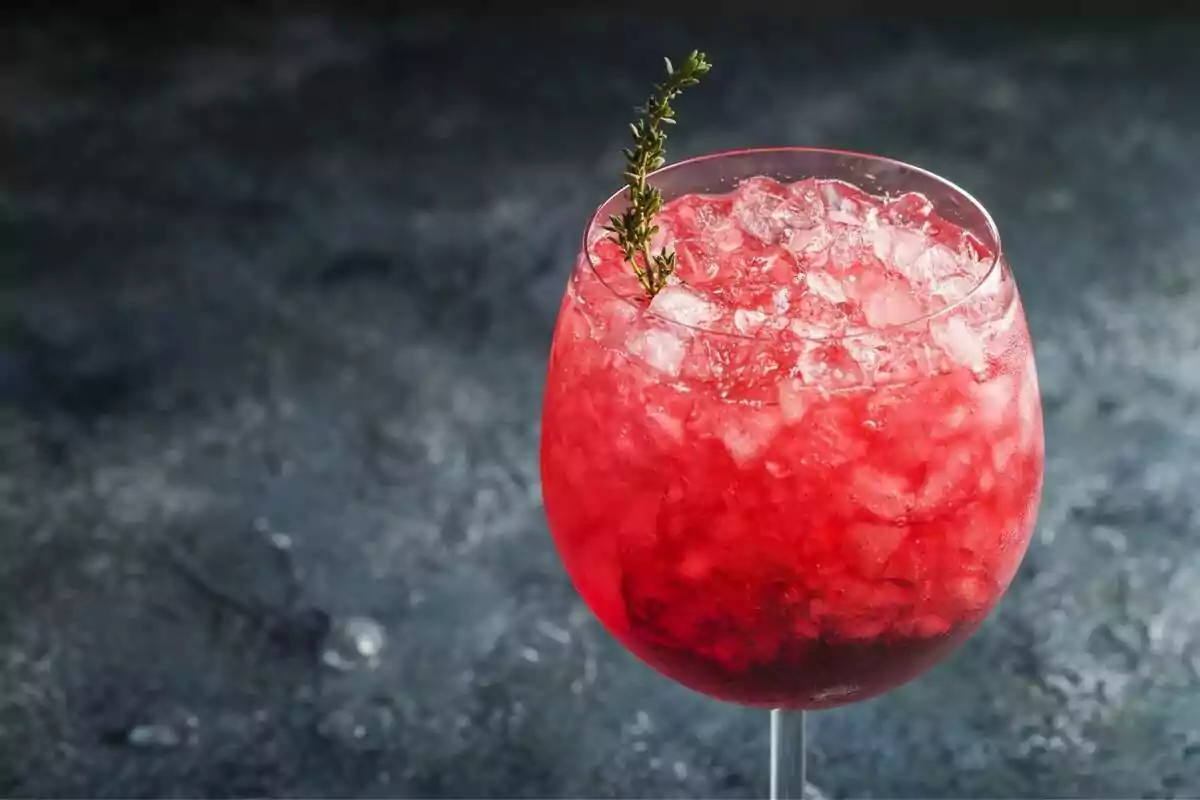 Copa de cóctel rojo con hielo picado y una ramita de hierba sobre una superficie oscura.