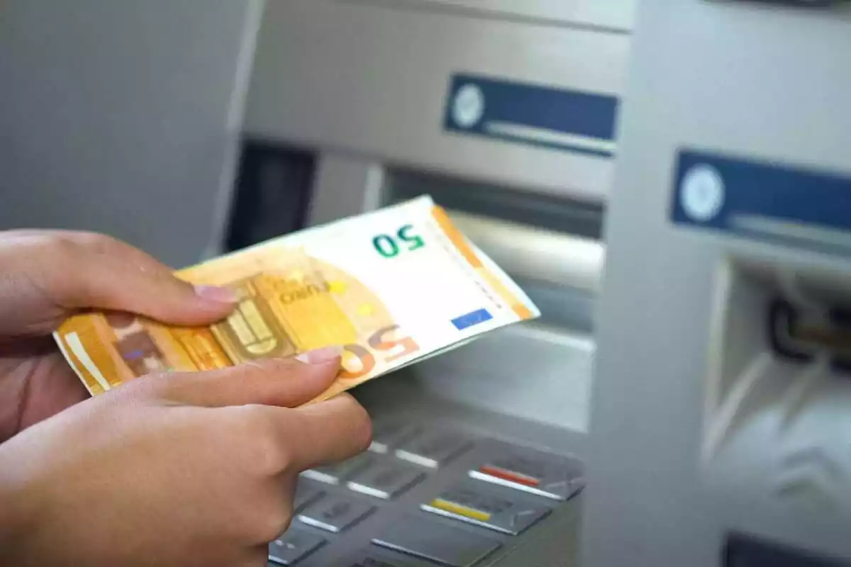 Dos manos de una persona sujetando billetes de 50 euros delante de un cajero automático