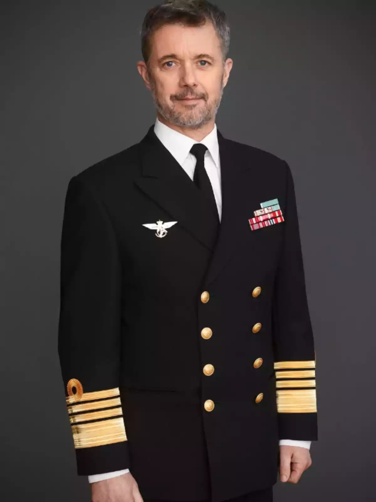 Hombre de mediana edad con barba y cabello corto vestido con uniforme militar oscuro con botones dorados y condecoraciones sobre un fondo gris Hombre de mediana edad con barba y cabello corto vestido con uniforme militar oscuro con botones dorados y condecoraciones sobre un fondo gris
