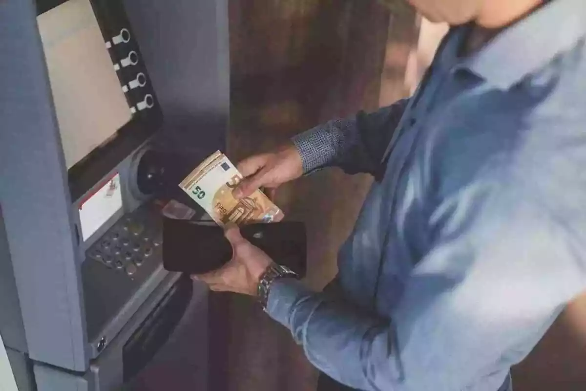 Hombre delante de un cajero automático con una cartera y billetes en las manos