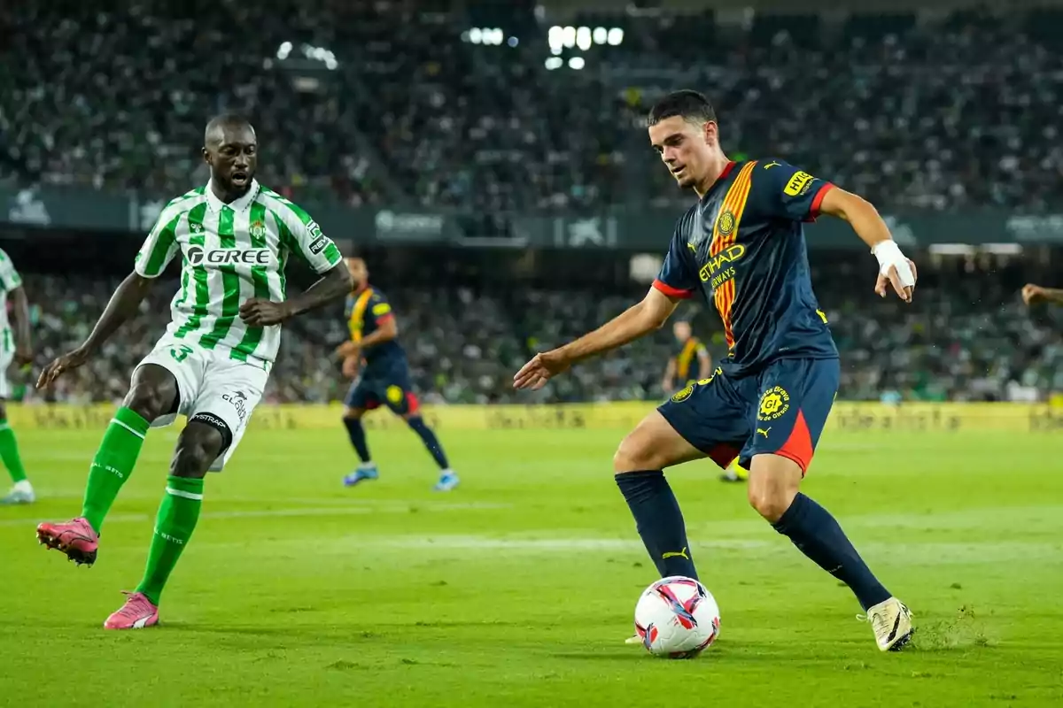 Dos jugadores de fútbol compiten por el balón en un partido, uno con uniforme verde y blanco y el otro con uniforme azul oscuro con detalles en rojo y amarillo. Dos jugadores de fútbol compiten por el balón en un partido, uno con uniforme verde y blanco y el otro con uniforme azul oscuro con detalles en rojo y amarillo.