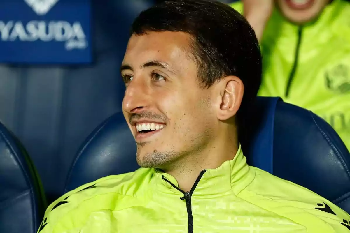 Un hombre sonriente con una chaqueta deportiva amarilla sentado en un asiento azul. Un hombre sonriente con una chaqueta deportiva amarilla sentado en un asiento azul.