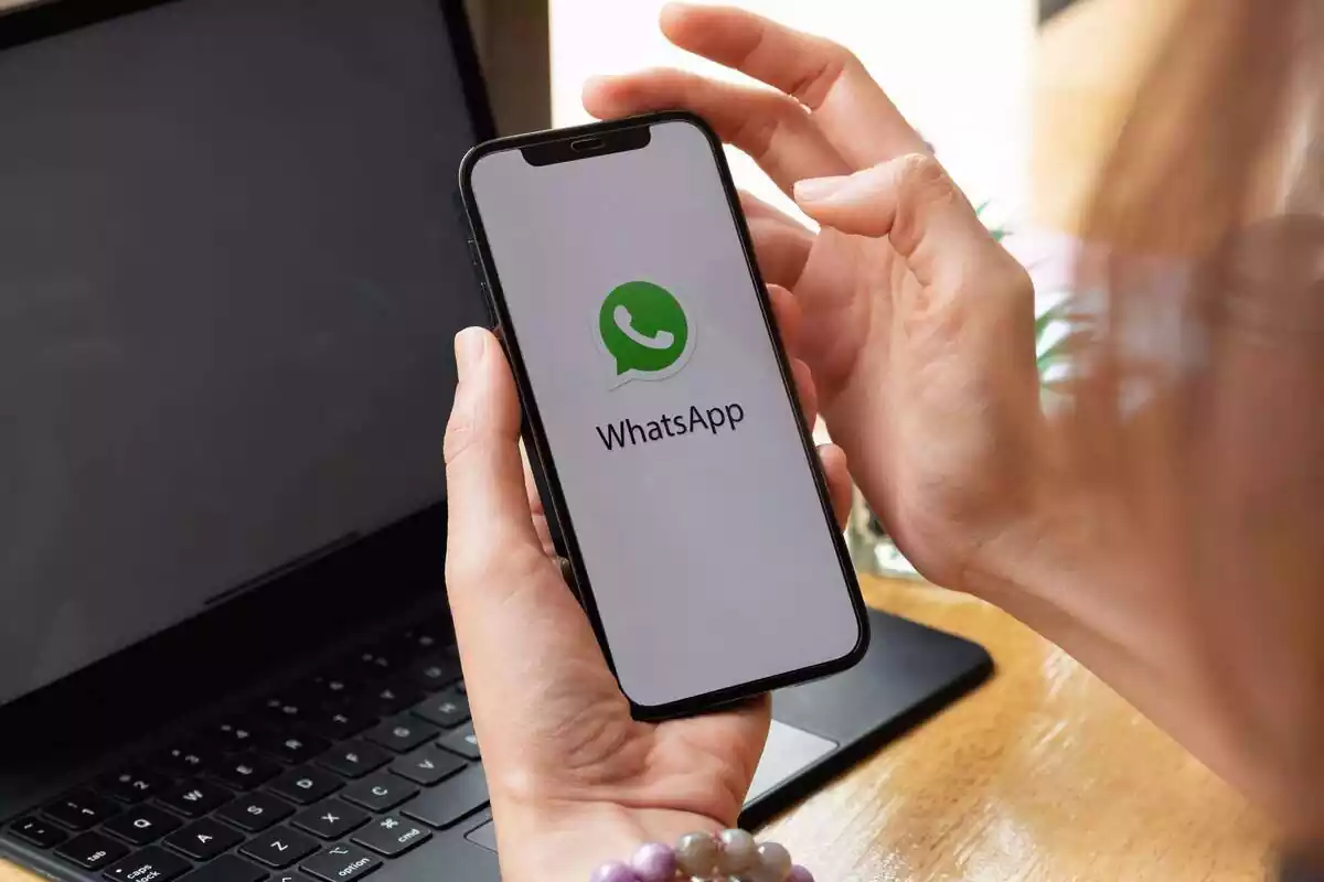 Manos de una mujer sosteniendo un móvil con el logo en medio de WhatsApp, de fondo un ordenador