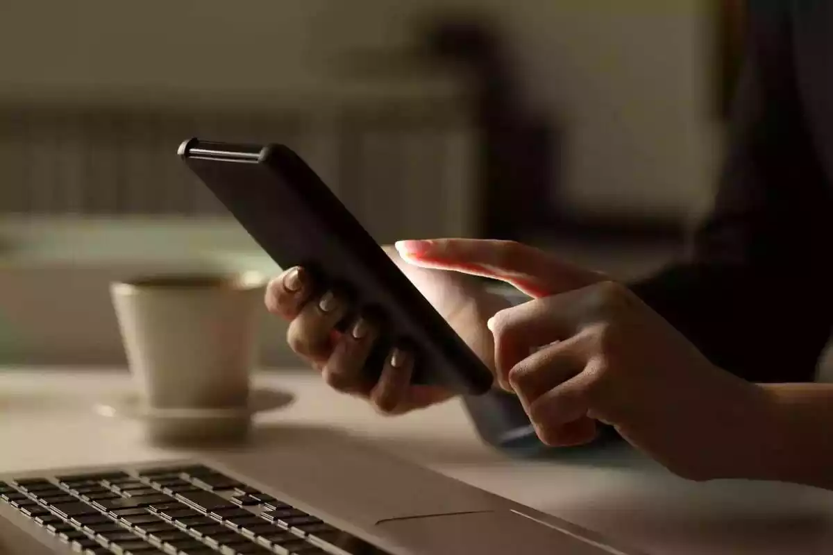 Una persona usando un móvil con un ordenador y una taza de café al lado