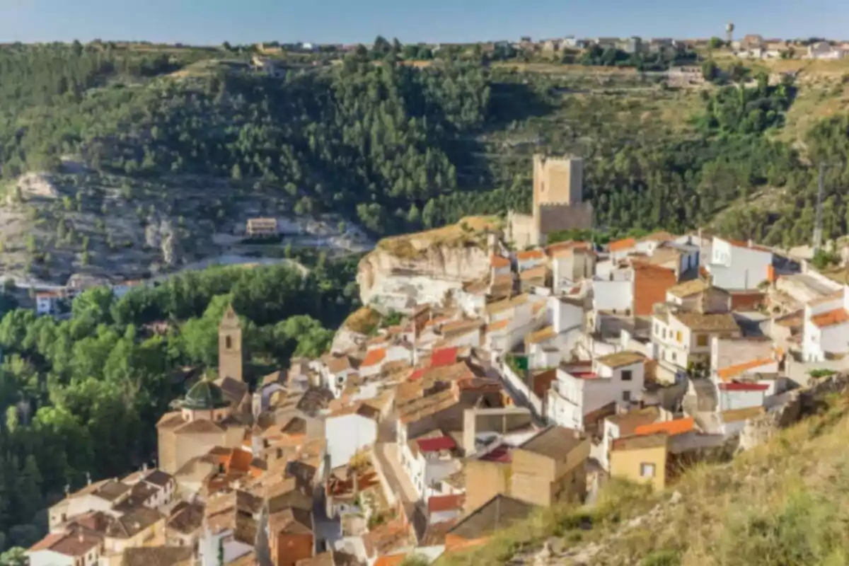 Vista panorámica de un pintoresco pueblo con casas de tejados rojos y una torre medieval, rodeado de colinas verdes y un paisaje montañoso.