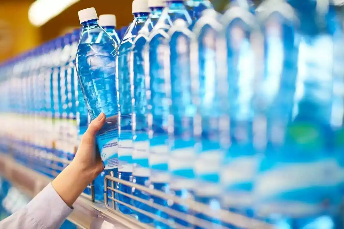 Una mano tomando una botella de agua de un estante lleno de botellas similares.