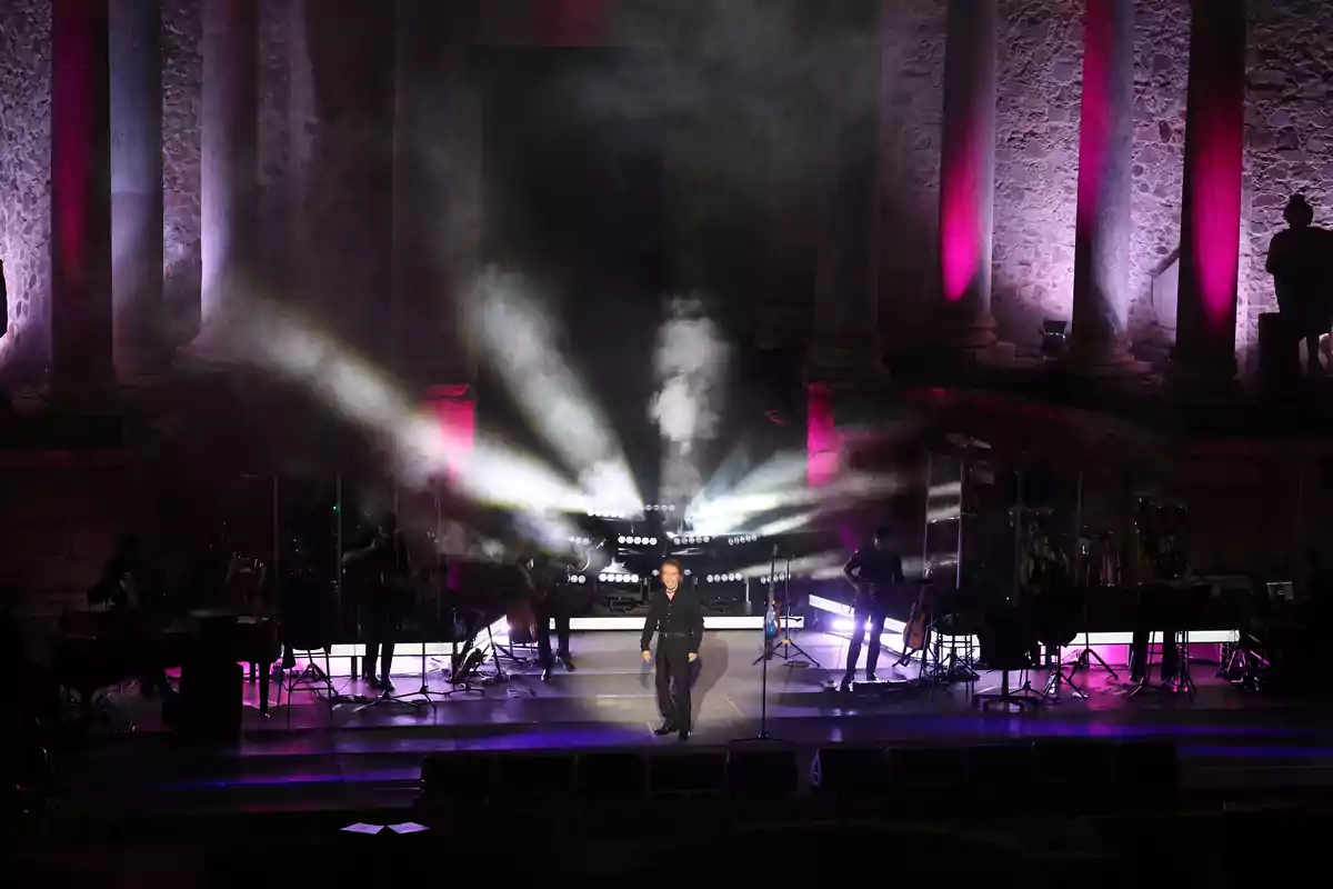 Raphael se presenta en un escenario iluminado con luces y humo, acompañado de músicos y rodeado de columnas antiguas iluminadas en tonos morados. Raphael se presenta en un escenario iluminado con luces y humo, acompañado de músicos y rodeado de columnas antiguas iluminadas en tonos morados.