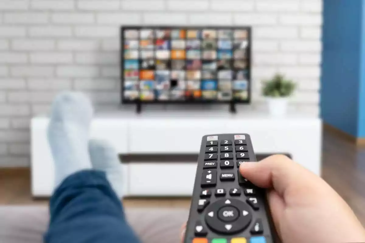 Persona relajada en un sofá usando un control remoto para cambiar canales en un televisor moderno.