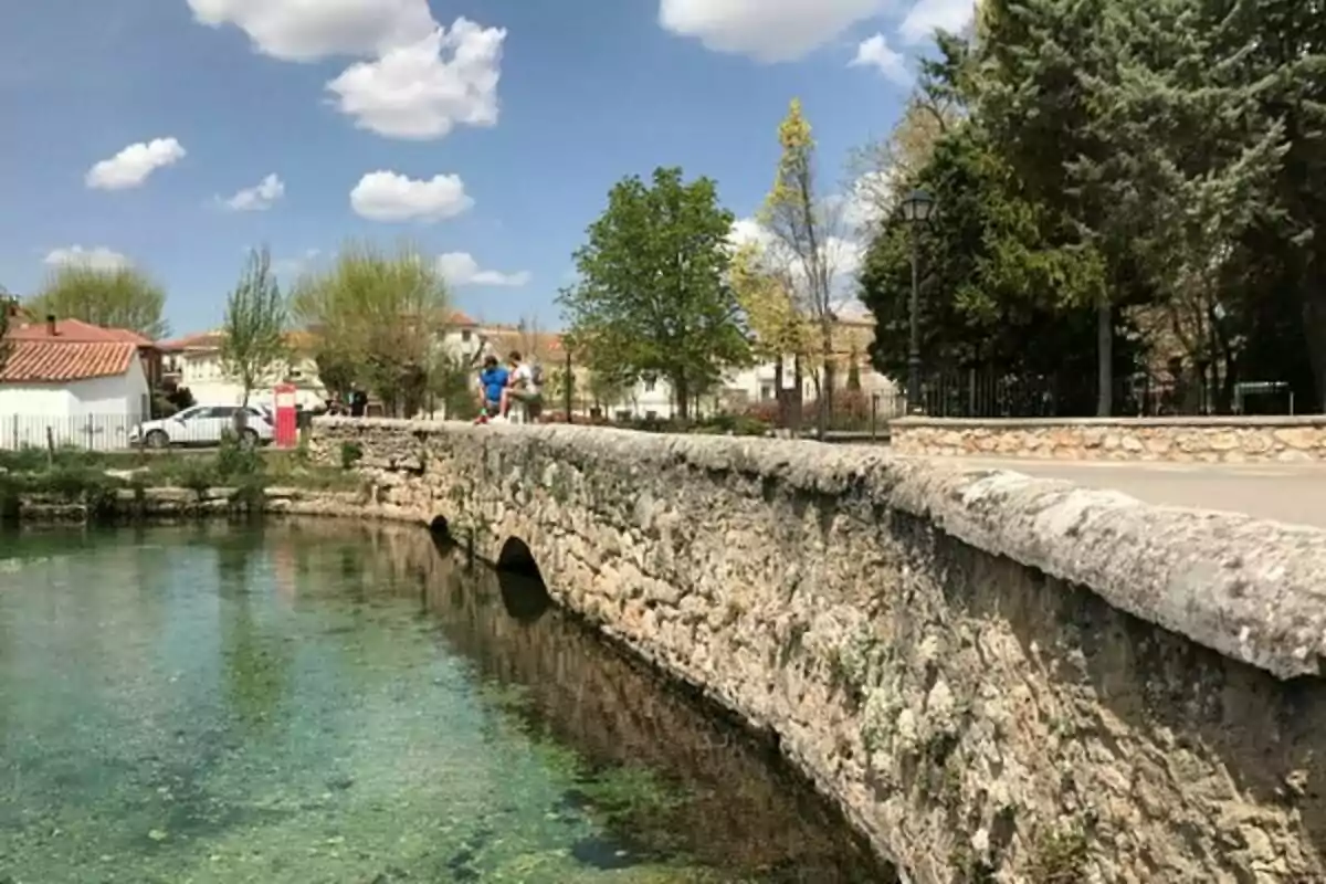 Un puente de piedra sobre un río con agua cristalina, rodeado de árboles y casas bajo un cielo azul con nubes.